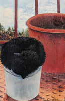 A canvas print featuring a black kitten asleep in a metal bucket, with a red background and a blurred garden in the background.